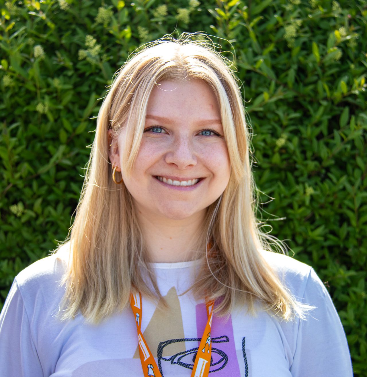 Trude smiler bredt. Hun har blondt rett hår til skuldrene og en hvit t-skjorte med abstrakt lilla trykk foran.