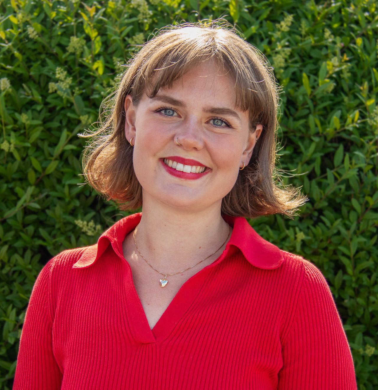Nora smiler bredt stående foran en grønn busk. Hun har skulderlangt brundt hår med lugg, rød leppestift og rød kortermet skjorte.