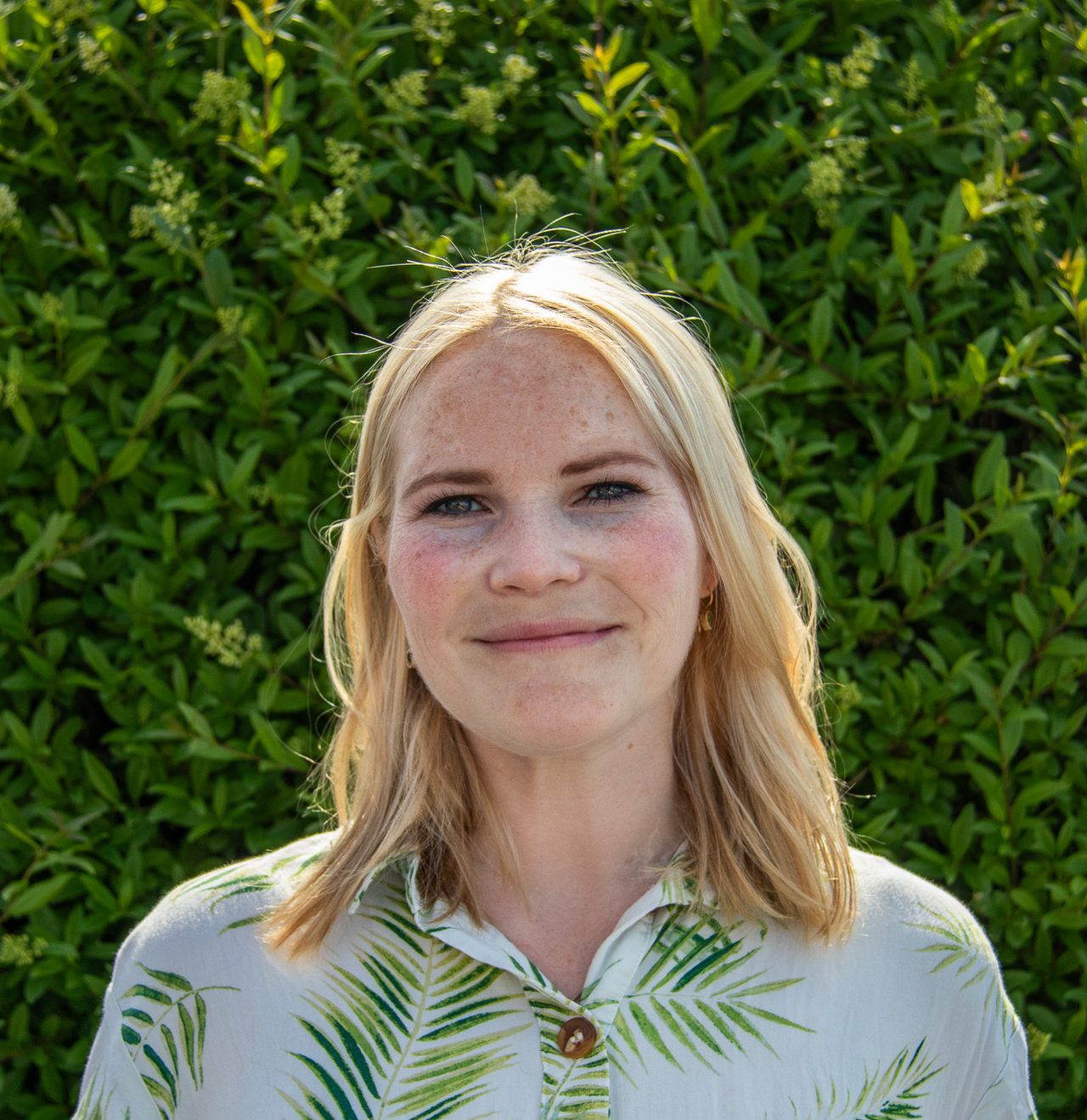 Hanne står foran en grønn busk. Hun har på se en kortermet hvit skjorte med grønne blader på, og blondt bølgete hår til skuldrene.