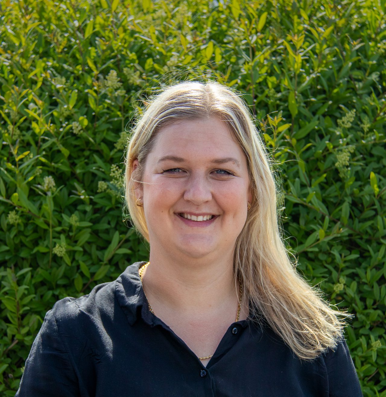 Ingvild smiler bredt mot kamera. Hun har langt blondt hår lagt til siden over den ene skulderen og har på seg en svart skjorte.