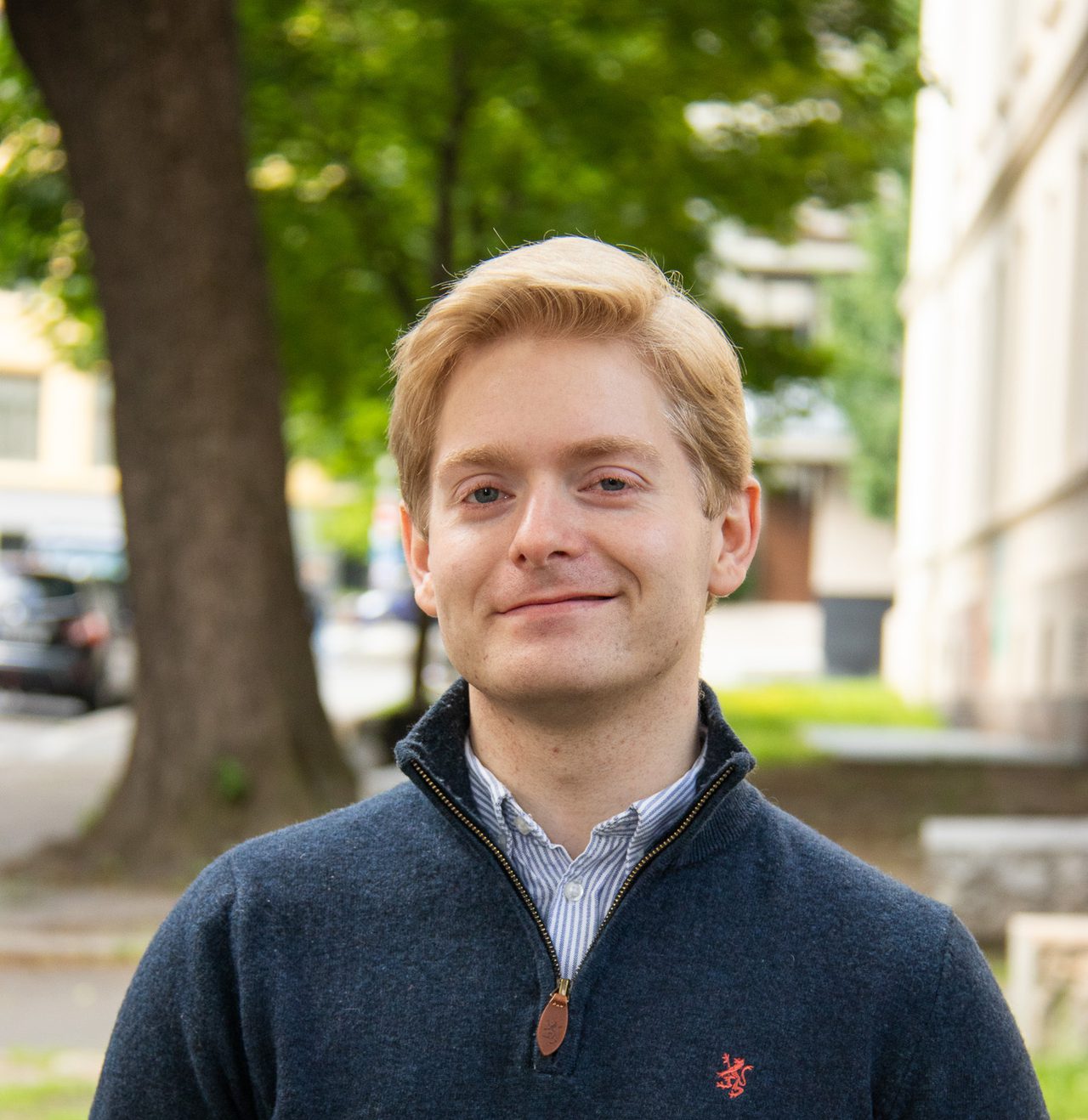 Amund står og smiler i sola. Han er veldig lys i huden og har kort lyst hår, lyse øyenbryn og lyse øyevipper. På seg har han en mørkeblå genser.