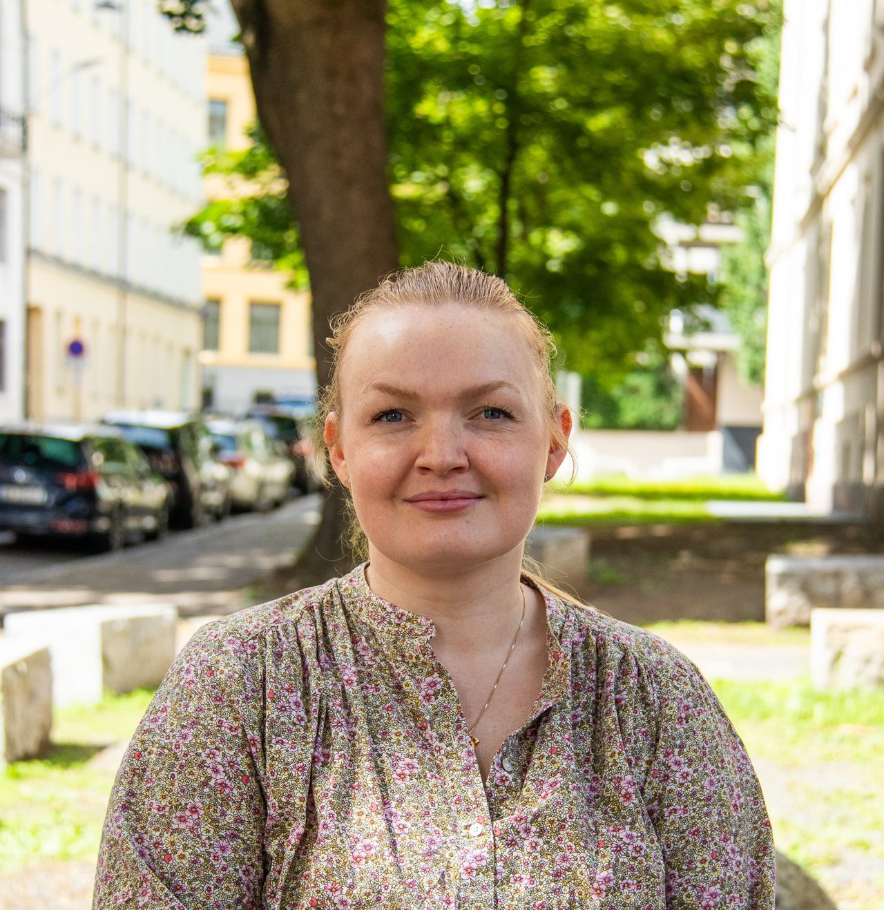 Emilie står og smiler ute i sola med det blonde håret oppsatt i en hestehale. Hun er lys i huden, har på sen en blomstrete skjorte og ser rett mot kameraet mens hun holder hendene sammen foran seg.