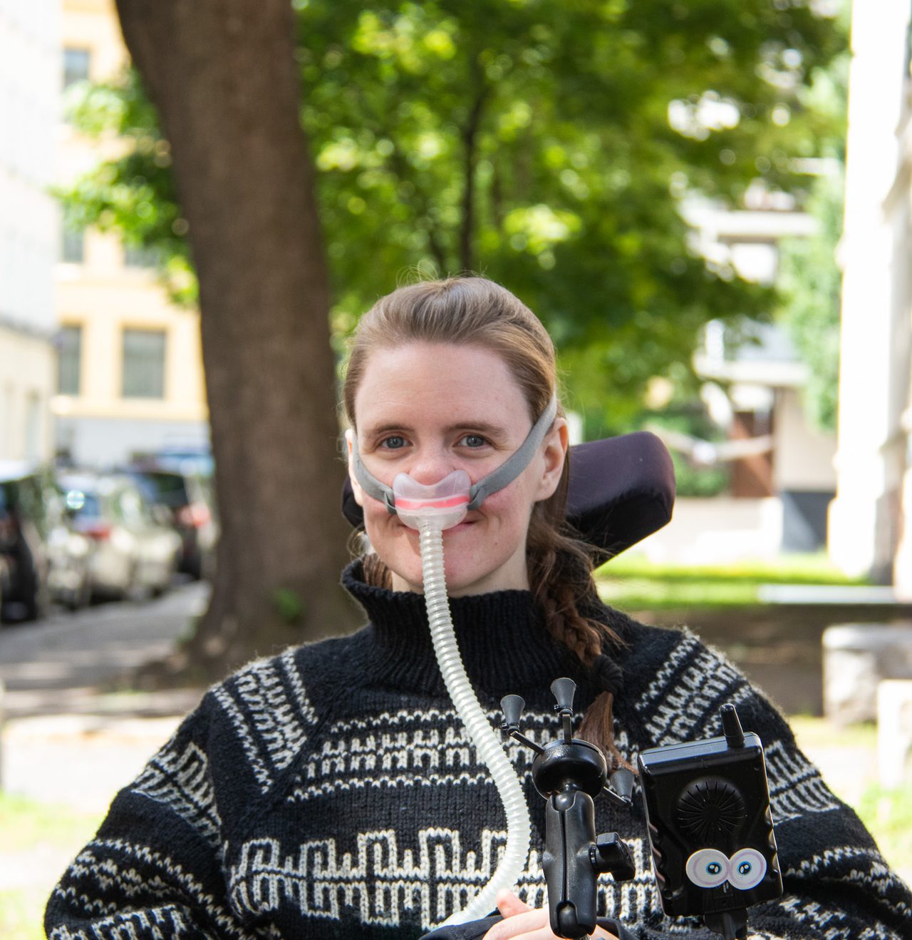 Ingrid sitter i en elektrisk rullestol ute på et fortau. Hun har en svart og hvit mønstret genser, mørkt hår i en hestehale og lys hud. Til nesen har hun koblet en respirator, med en slange som går ned til en pustemaskin. Den er festet rundt hodet hennes med en gummistropp.