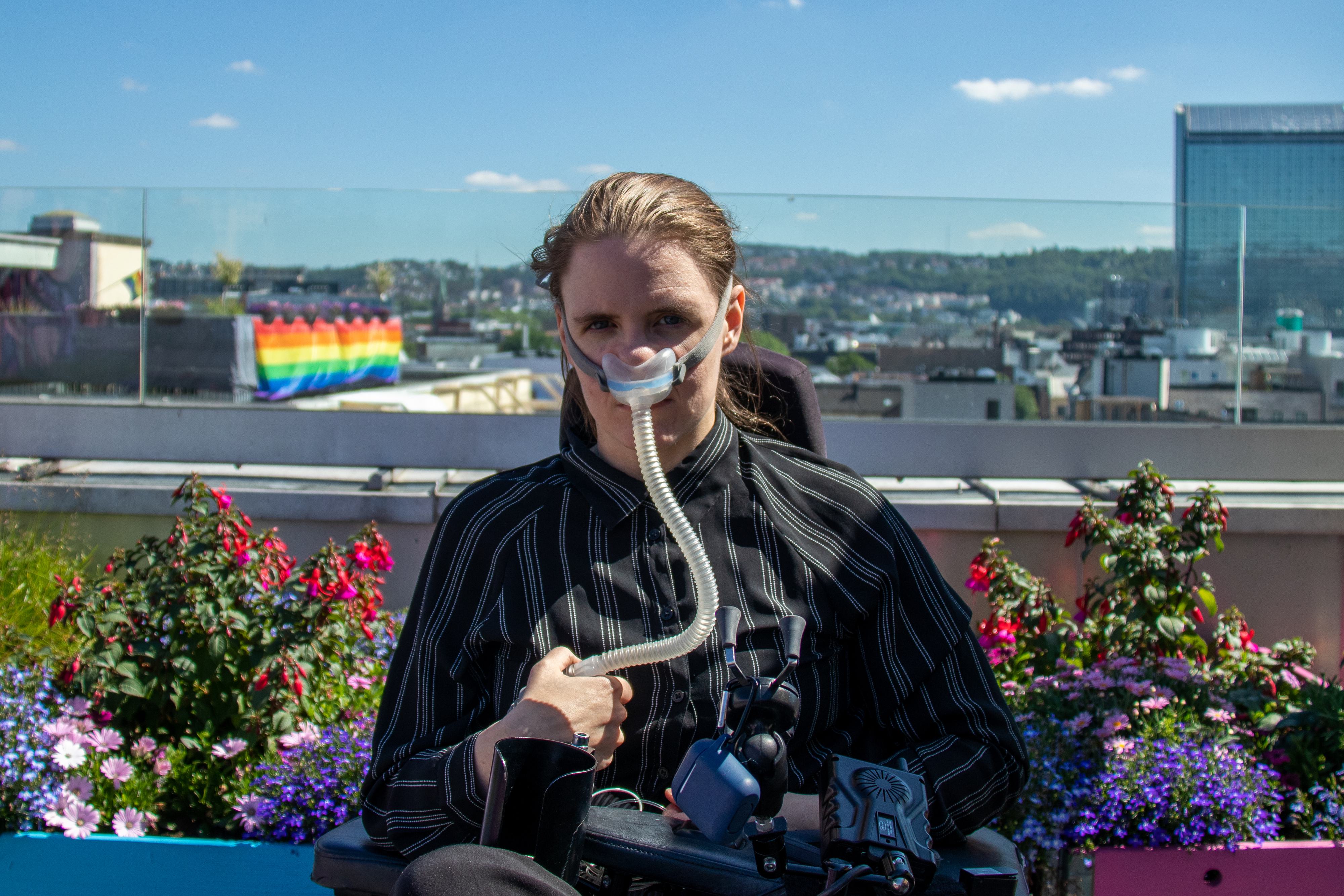 Ingrid sitter i en elektrisk rullestol oppe på en terrasse med utsikt over en by. De ser alvorlig inn i kamera. De har en svart og hvit stripete skjorte, mørkt hår i en hestehale og lys hud. Til nesen har de koblet en respirator, med en slange som går ned til en pustemaskin. Den er festet rundt hodet med en gummistropp.