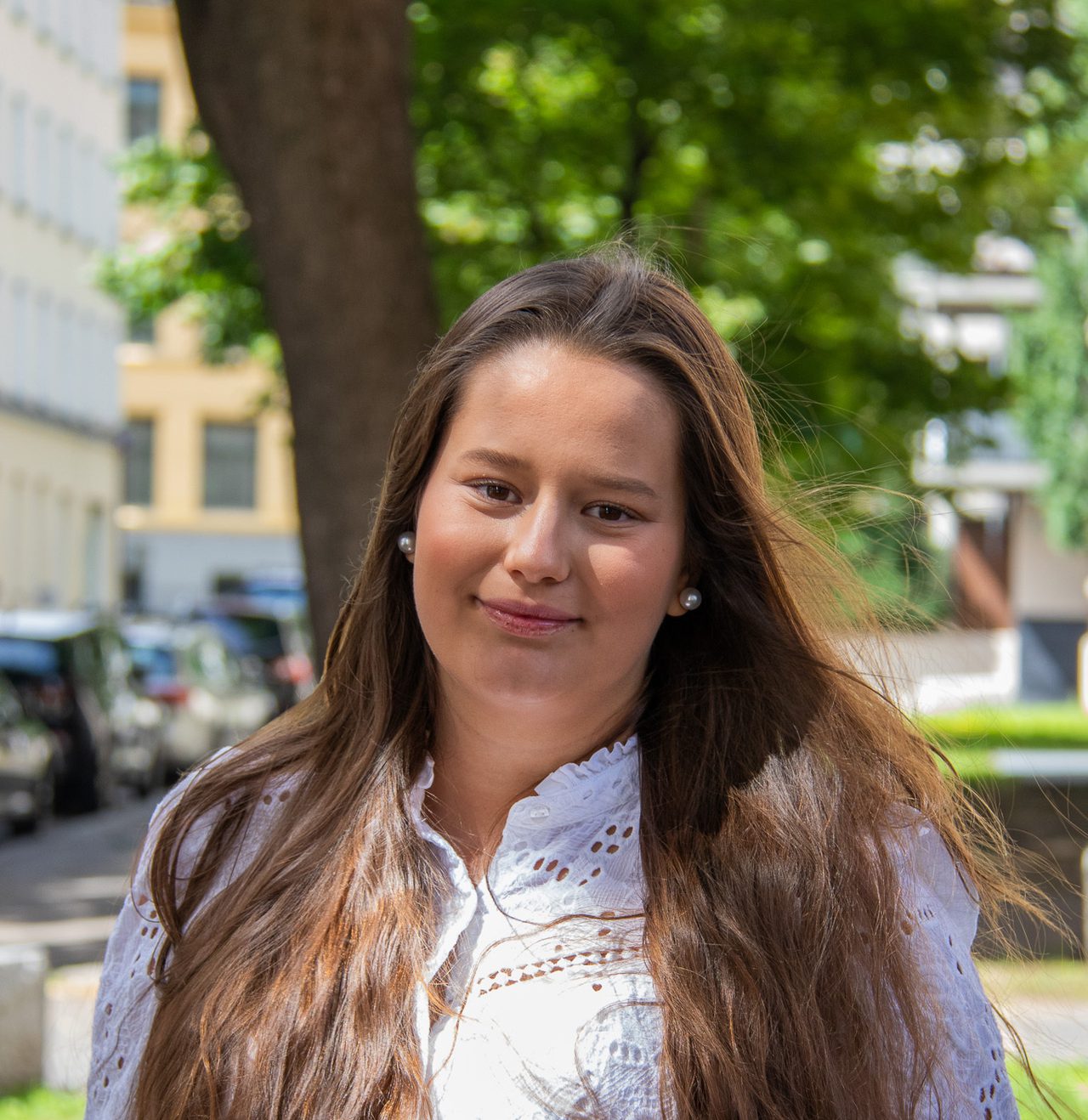 Thea har langt brunt hår, er lys i huden og har på seg hvit langermet skjorte. Hun står ute i sola på et fortau med hendene i lommene og et lite smil mot kameraet.