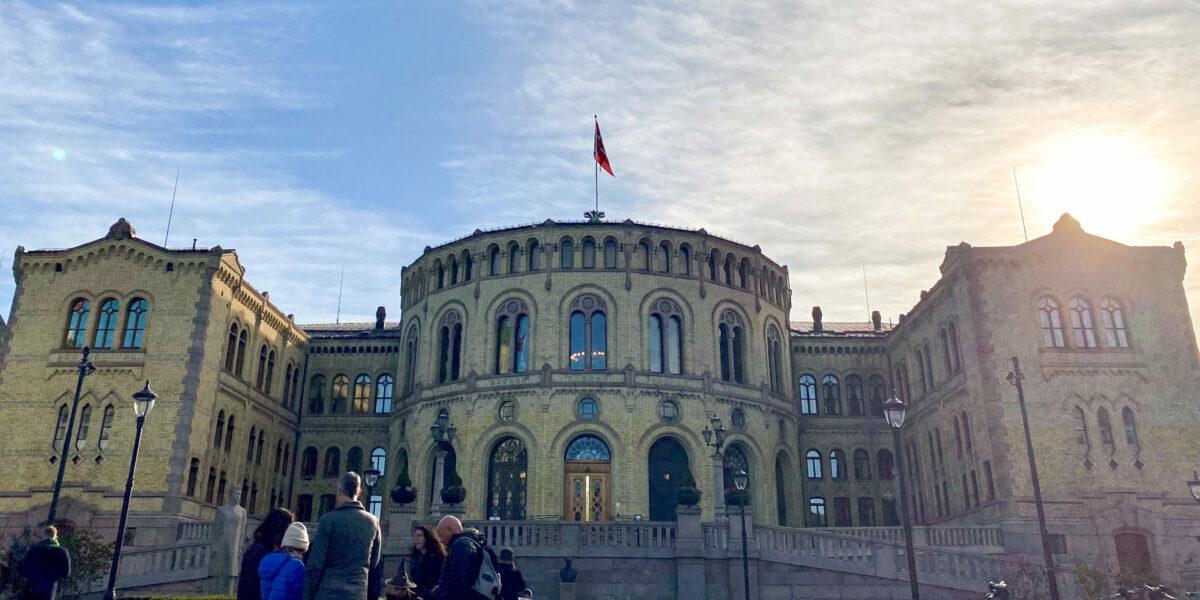 Stortinget i solskinn med 5 mennesker foran bygningen.