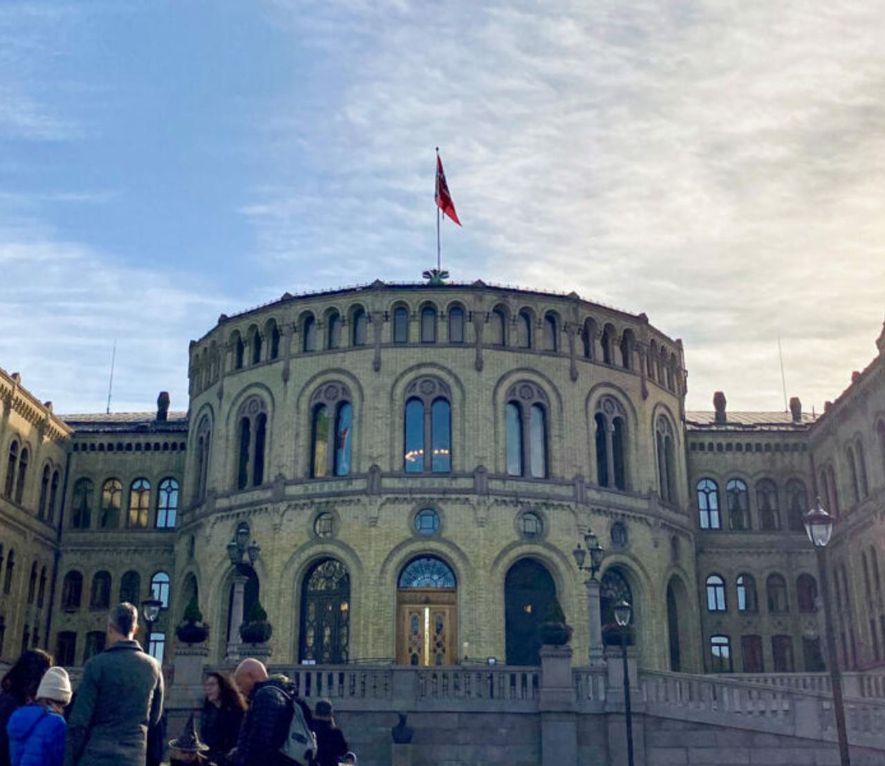 Stortinget i solskinn med 5 mennesker foran bygningen.