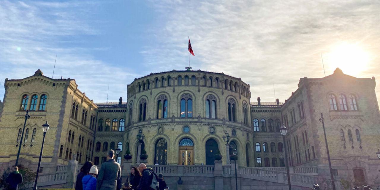 Stortinget i solskinn med 5 mennesker foran bygningen.