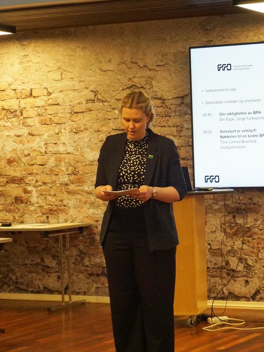 Siri står oppreist og offisielt og leser fra talekortene hun har i hendene. På seg har hun en svart blazer, svart bukse og rundt hodet har hun en mikrofon.