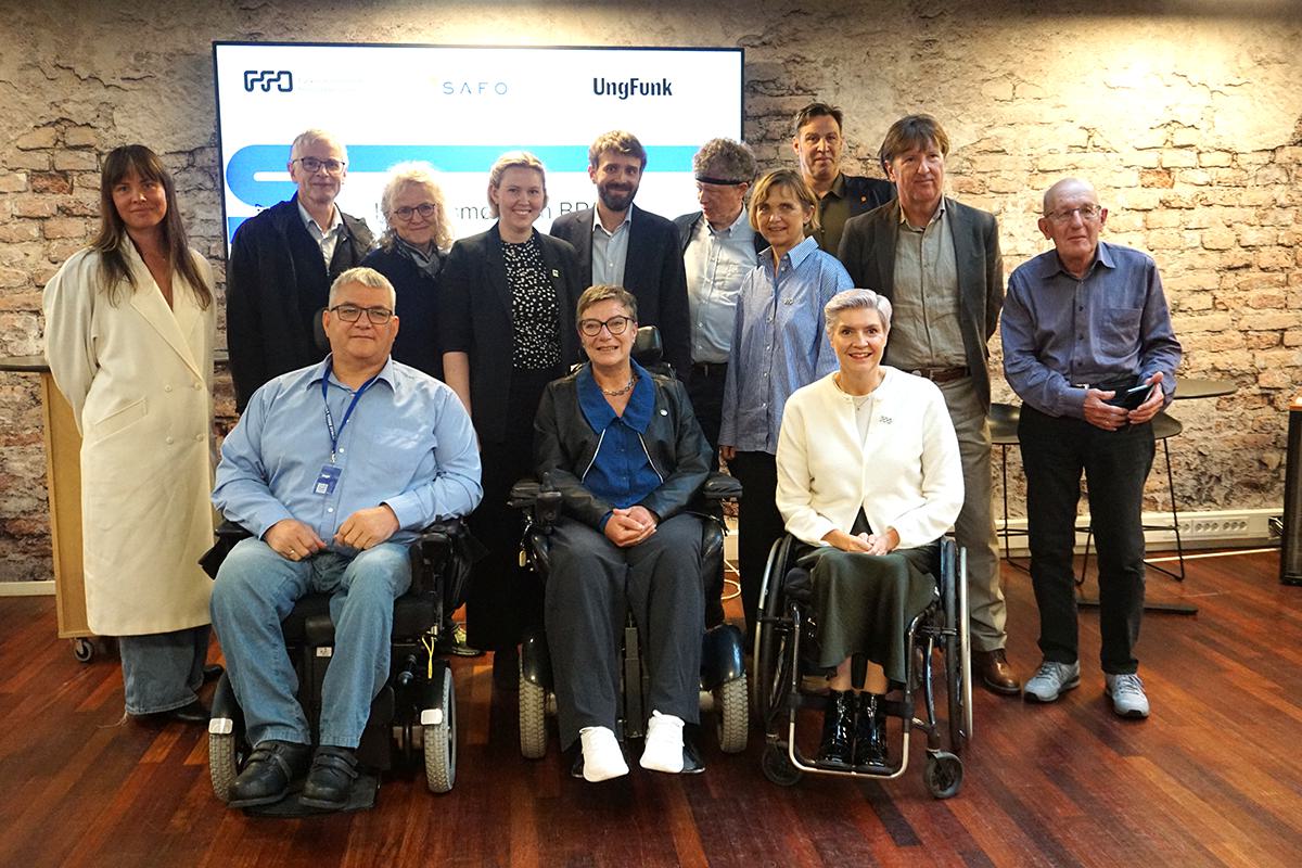 Rundt Jan Christian Vestre står mange representanter fra orgnaisasjonene og smiler. Det står en rekke bak og det sitter tre mennesker foran i rullestol.