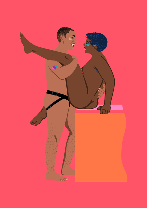 En person med helt kort hår står og holder tett rundt en kvinne. Personen som står har en strap-on på seg, og et hjerte på skulderen i Pride-farger. Personen smiler stort til kvinnen. Kvinnen sitter på kanten av et møbel, hun har mørk hud, små pupper, kort mørkt hår og store briller. Hun har det ene benet over skulderen til sin partner, og det andre rundt partnerens hofter. Også hun smiler.