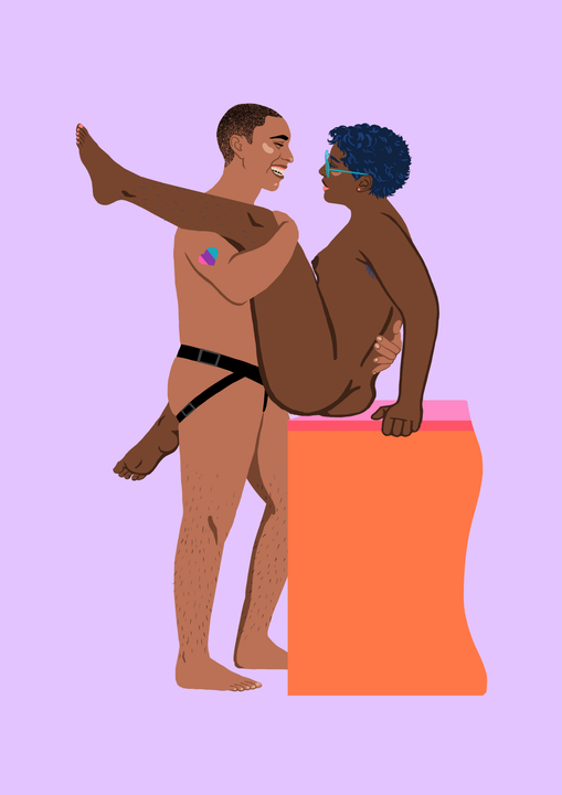 En person med helt kort hår står og holder tett rundt en kvinne. Personen som står har en strap-on på seg, og et hjerte på skulderen i Pride-farger. Personen smiler stort til kvinnen. Kvinnen sitter på kanten av et møbel, hun har mørk hud, små pupper, kort mørkt hår og store briller. Hun har det ene benet over skulderen til sin partner, og det andre rundt partnerens hofter. Også hun smiler.