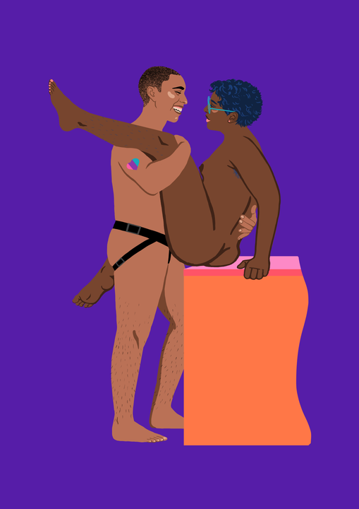 En person med helt kort hår står og holder tett rundt en kvinne. Personen som står har en strap-on på seg, og et hjerte på skulderen i Pride-farger. Personen smiler stort til kvinnen. Kvinnen sitter på kanten av et møbel, hun har mørk hud, små pupper, kort mørkt hår og store briller. Hun har det ene benet over skulderen til sin partner, og det andre rundt partnerens hofter. Også hun smiler.