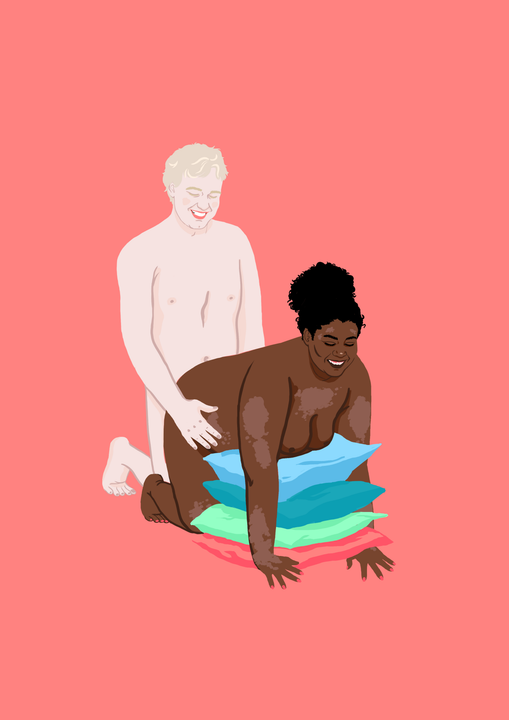 En mørk, kraftig kvinne står på knærne foran en veldig lys, slank mann. Kvinnen lener overkroppen over en rad med puter. Hun har psoriasis flekker ulike steder på kroppen, og smiler. Mannen står på knærne bak kvinnen og holder kvinnen rundt hoftene. Også han smiler.