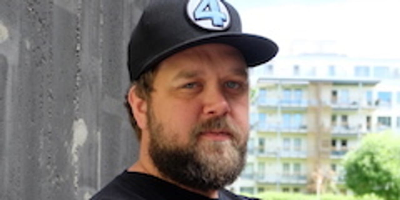 Ung mann ser inn i kamera. Han har blå øyne, mørkeblondt skjegg og lys hud. Han har på seg en svart caps og en svart t-skjorte.