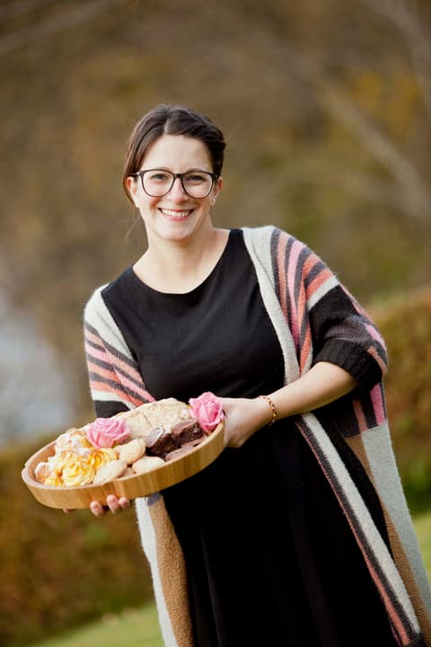 Ung kvinne står på en gressplen og ser inn i kamera og smiler. I hendene foran seg holder hun et fat med kaker og blomster. Hun har brunt hår som er satt opp, lys hud, mørke øyne og svarte briller. Hun har på seg en svart kjole og en strikket stripete jakke i rosa, hvit og brun.