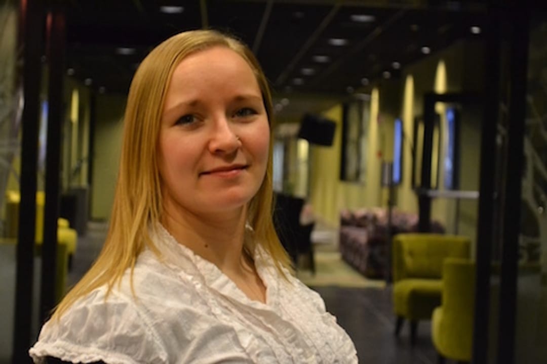 Unge kvinne ser inn i kamera og smiler. Hun har lyst langt hår og lys hud. Hun har på seg en hvit bluse.